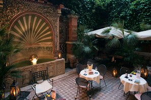 Un patio al aire libre con muros de piedra ornamentados, una fuente y mesas dispuestas para comer.