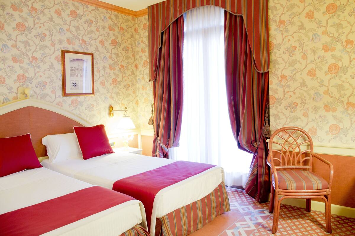 Ein Hotelzimmer mit zwei Betten, einem Holzstuhl und einem großen Fenster mit Vorhängen.