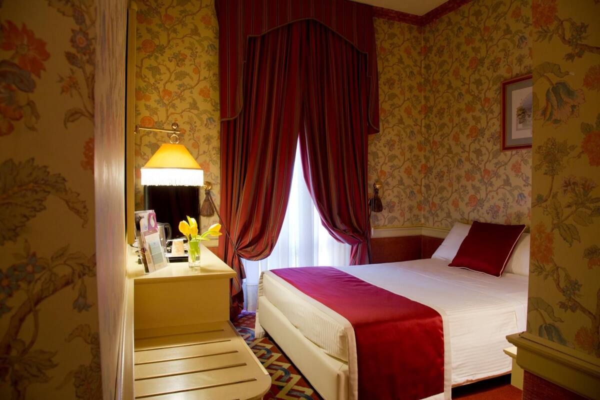 Ein Hotelzimmer mit Bett, Schreibtisch mit Lampe, Fenster mit Vorhängen und Blumenwallpaper.