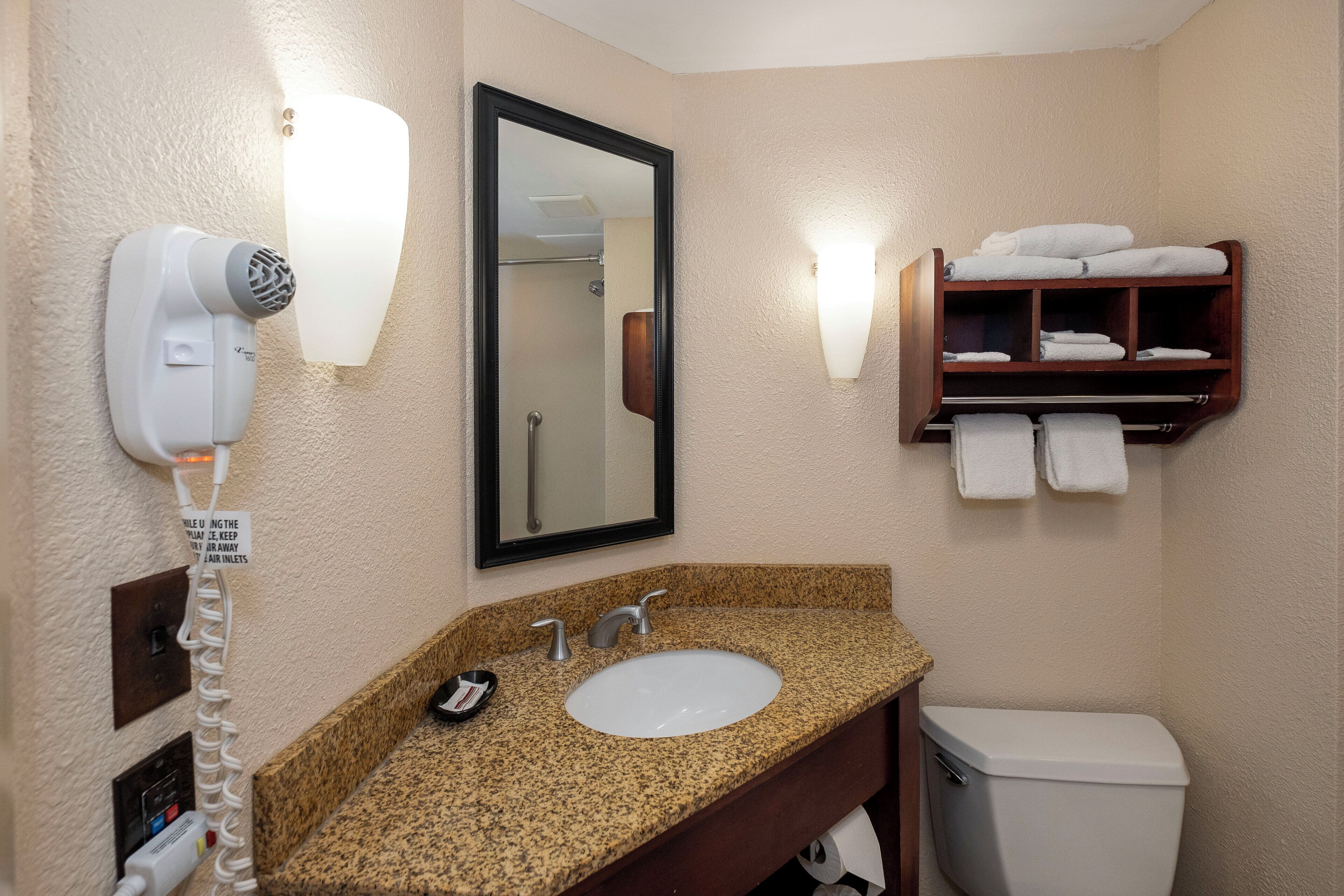 A hotel bathroom with a granite countertop sink, a mirror, a towel rack, and a hair dryer.