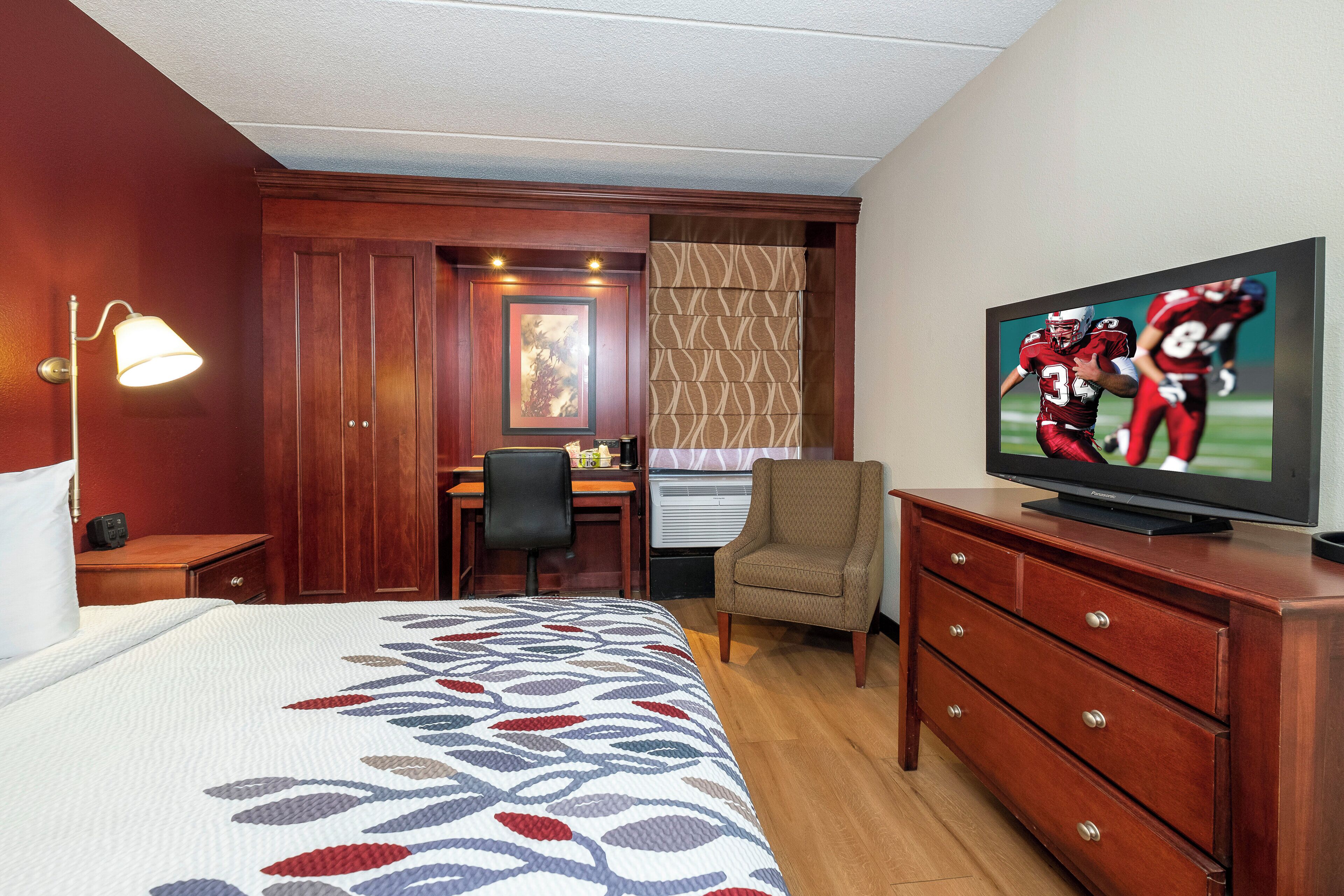Quarto de hotel com cama, escrivaninha, cadeira, televisão e cómoda.