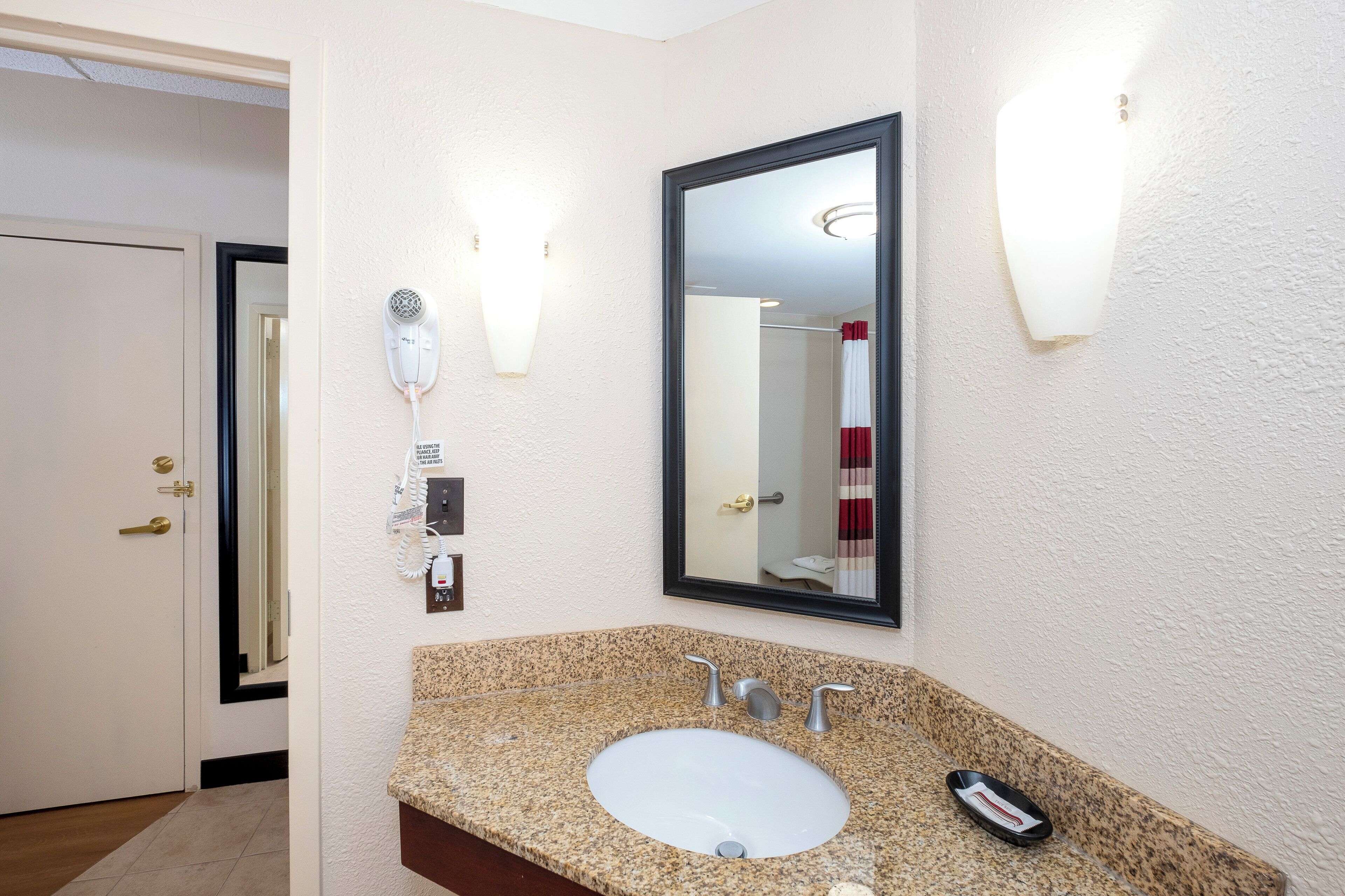 A bathroom with a granite countertop sink, a mirror, and a hairdryer mounted on the wall.