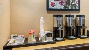A coffee station with hot water, regular coffee, and decaf coffee dispensers, a stack of white cups, condiments, and a framed picture of red flowers on the wall.