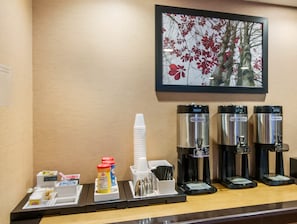 A coffee station with hot water, regular coffee, and decaf coffee dispensers, a stack of white cups, condiments, and a framed picture of red flowers on the wall.