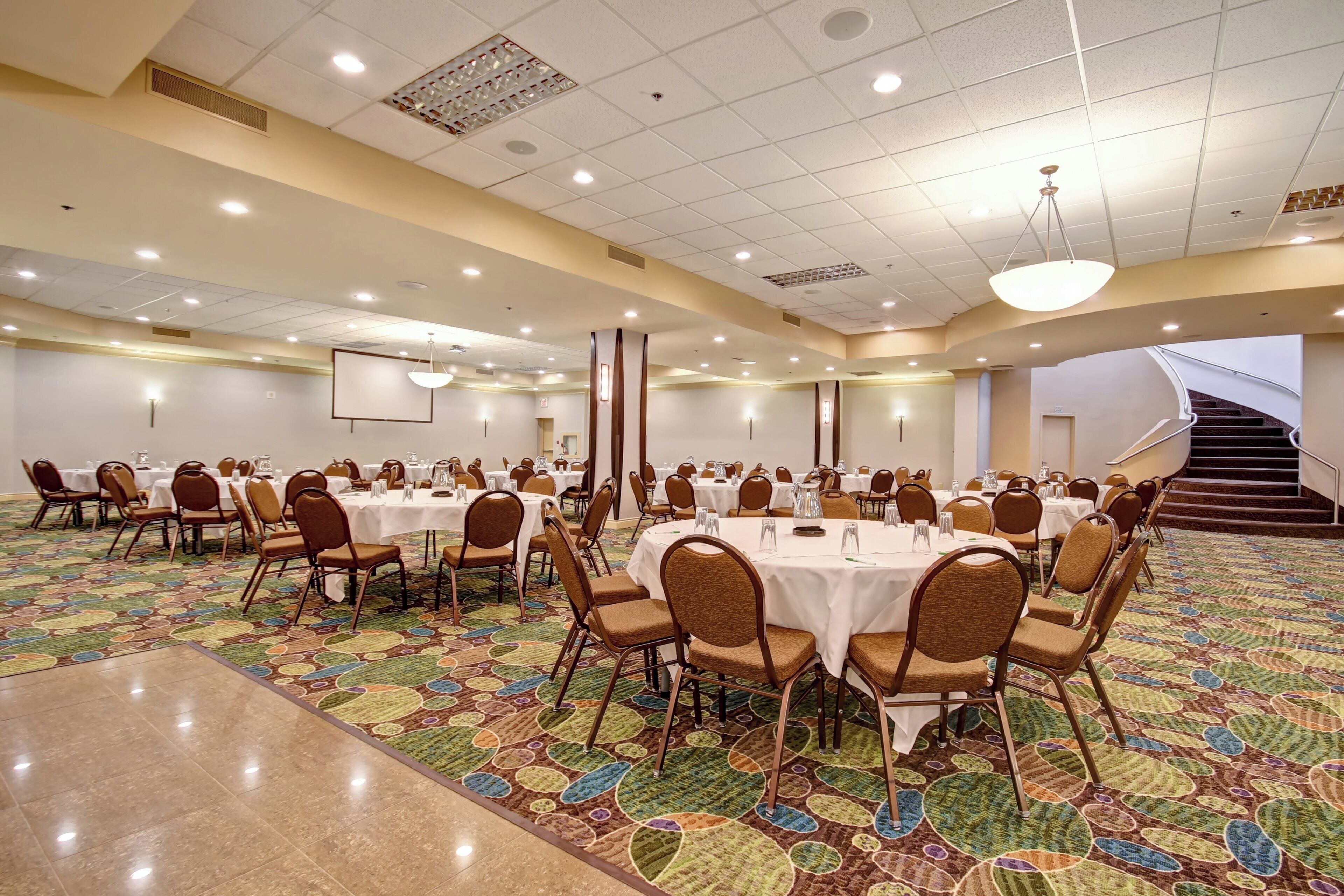 A large conference room with multiple tables and chairs, a projector screen, and a staircase leading to an upper level.
