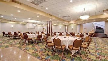 A large conference room with multiple tables and chairs, a projector screen, and a staircase leading to an upper level.