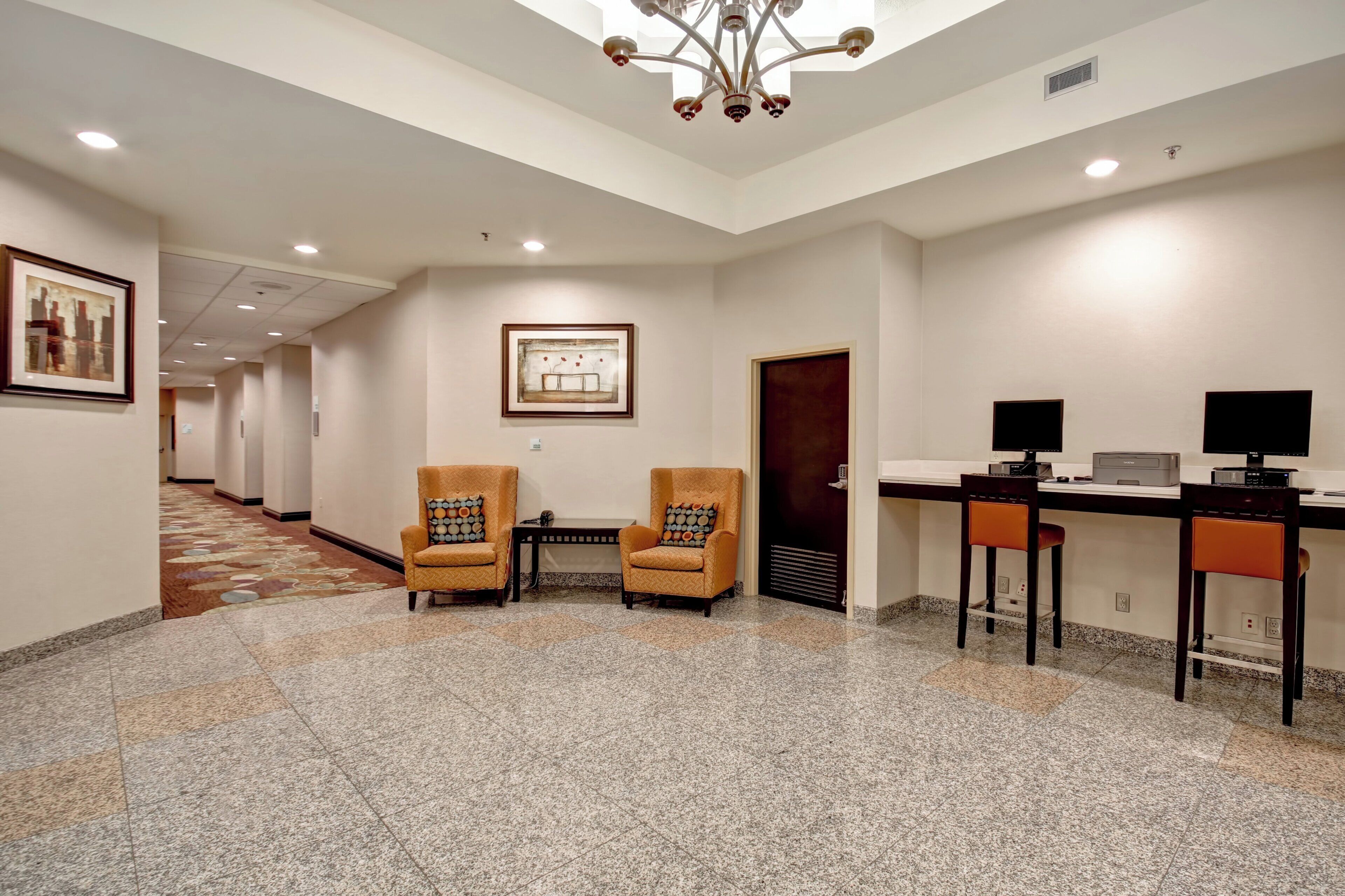 Um hall de hotel com piso carpetado, dois poltronas, uma mesa com computador e uma obra de arte emoldurada na parede.