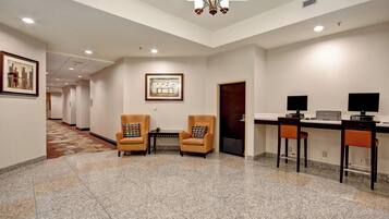 Um hall de hotel com piso carpetado, dois poltronas, uma mesa com computador e uma obra de arte emoldurada na parede.