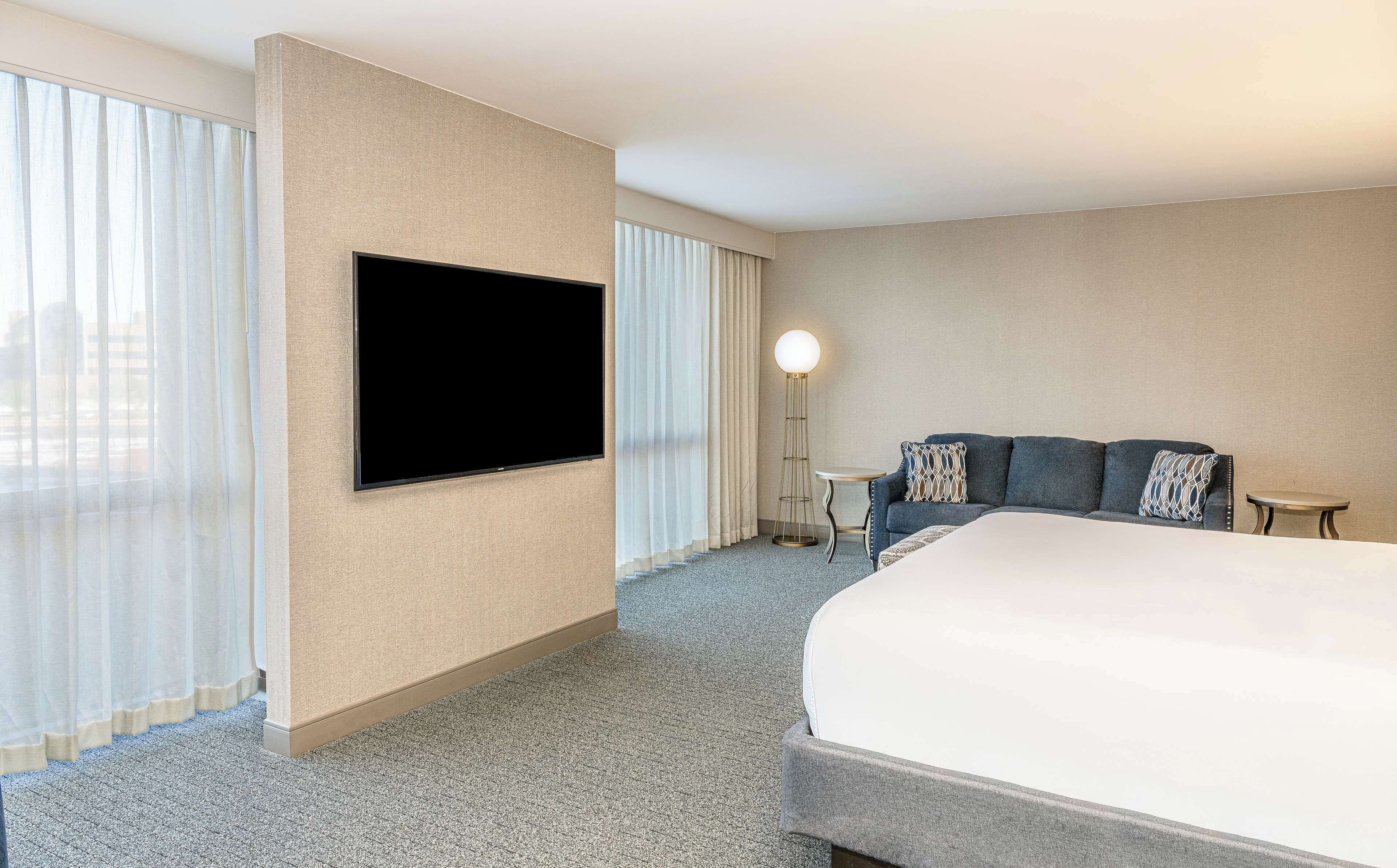 A hotel room with a large bed, a flat-screen TV mounted on the wall, a floor lamp, a sofa with patterned cushions, and a small table.
