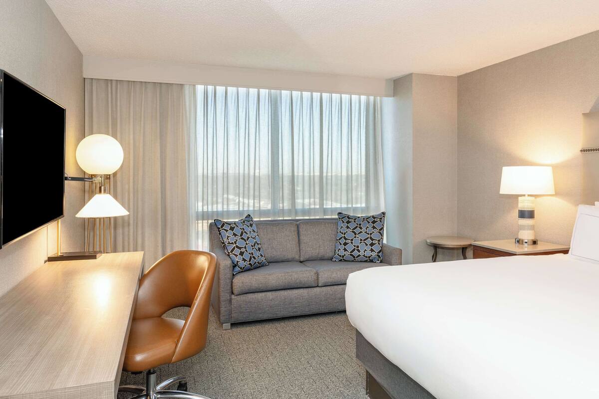 Quarto de hotel com cama, escrivaninha, cadeira, sofá e televisão.