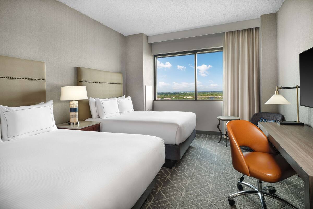 Quarto de hotel com duas camas, uma escrivaninha, uma cadeira, uma luminária e vista para a cidade.
