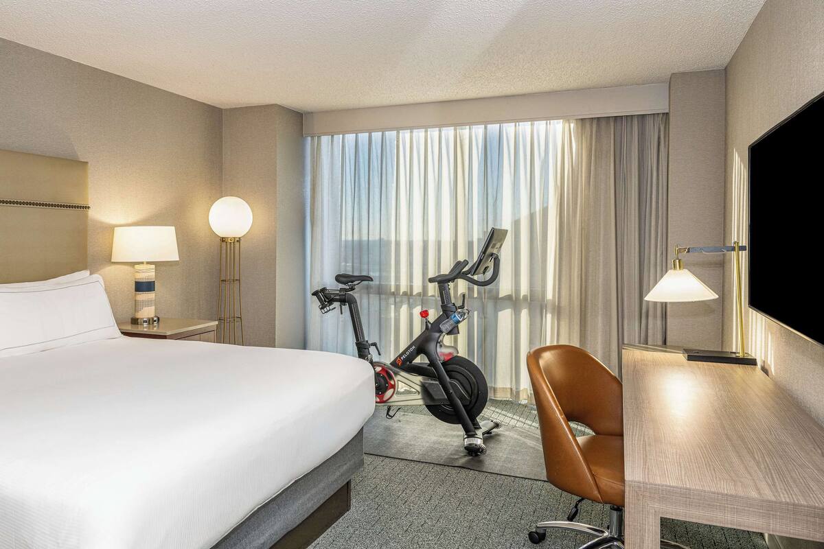 Quarto de hotel com uma cama grande, uma escrivaninha com cadeira, abajur, bicicleta e televisão.
