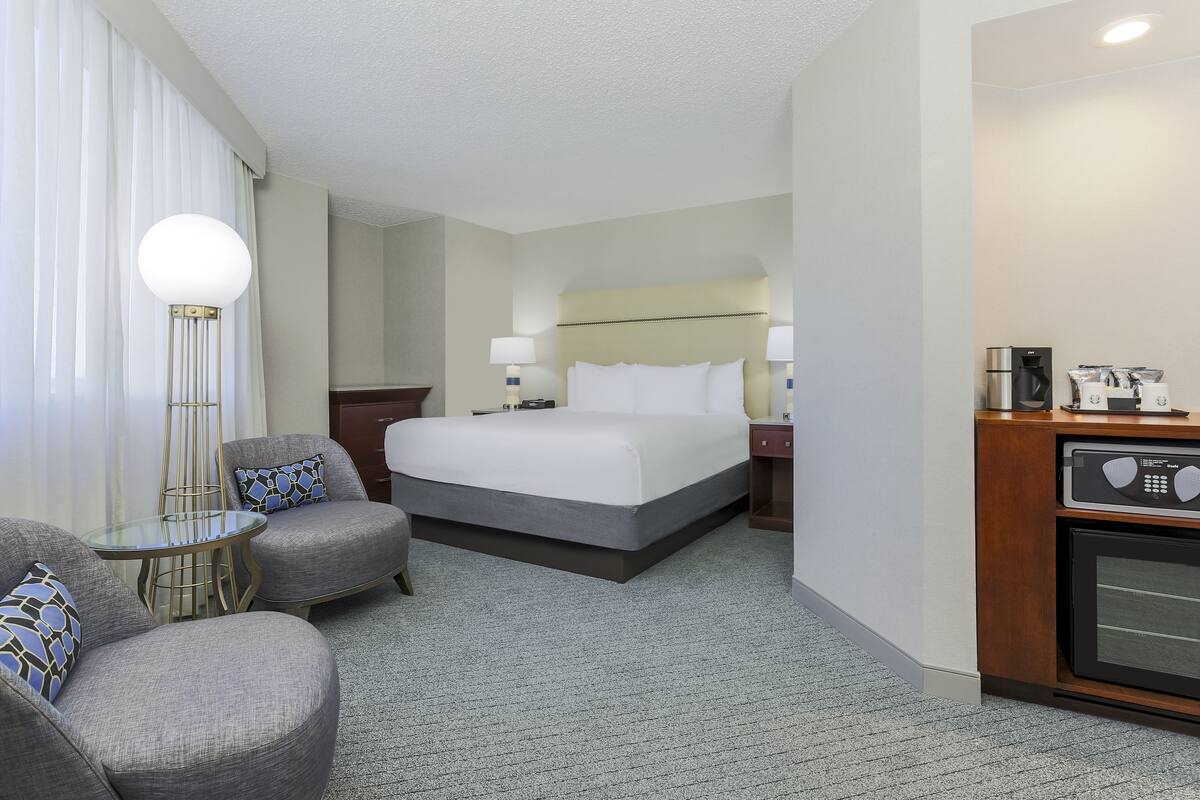Quarto de hotel com cama, duas poltronas, uma mesinha, luminária, cafeteira e micro-ondas.