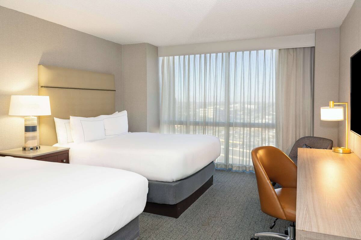 Quarto de hotel com duas camas, uma escrivaninha, uma cadeira, televisão e uma janela grande com cortinas.