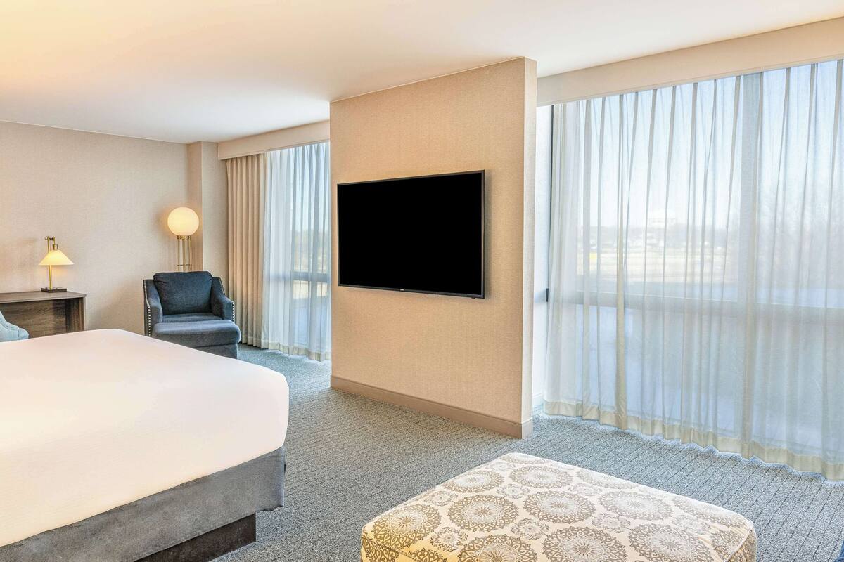 Quarto de hotel com uma cama grande, televisão de tela plana na parede, uma cadeira, uma luminária e janelas amplas com cortinas finas.