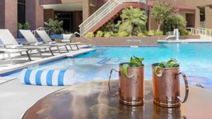 A poolside view with two copper mugs on a table, a pool, and a building with a red roof and brick walls.