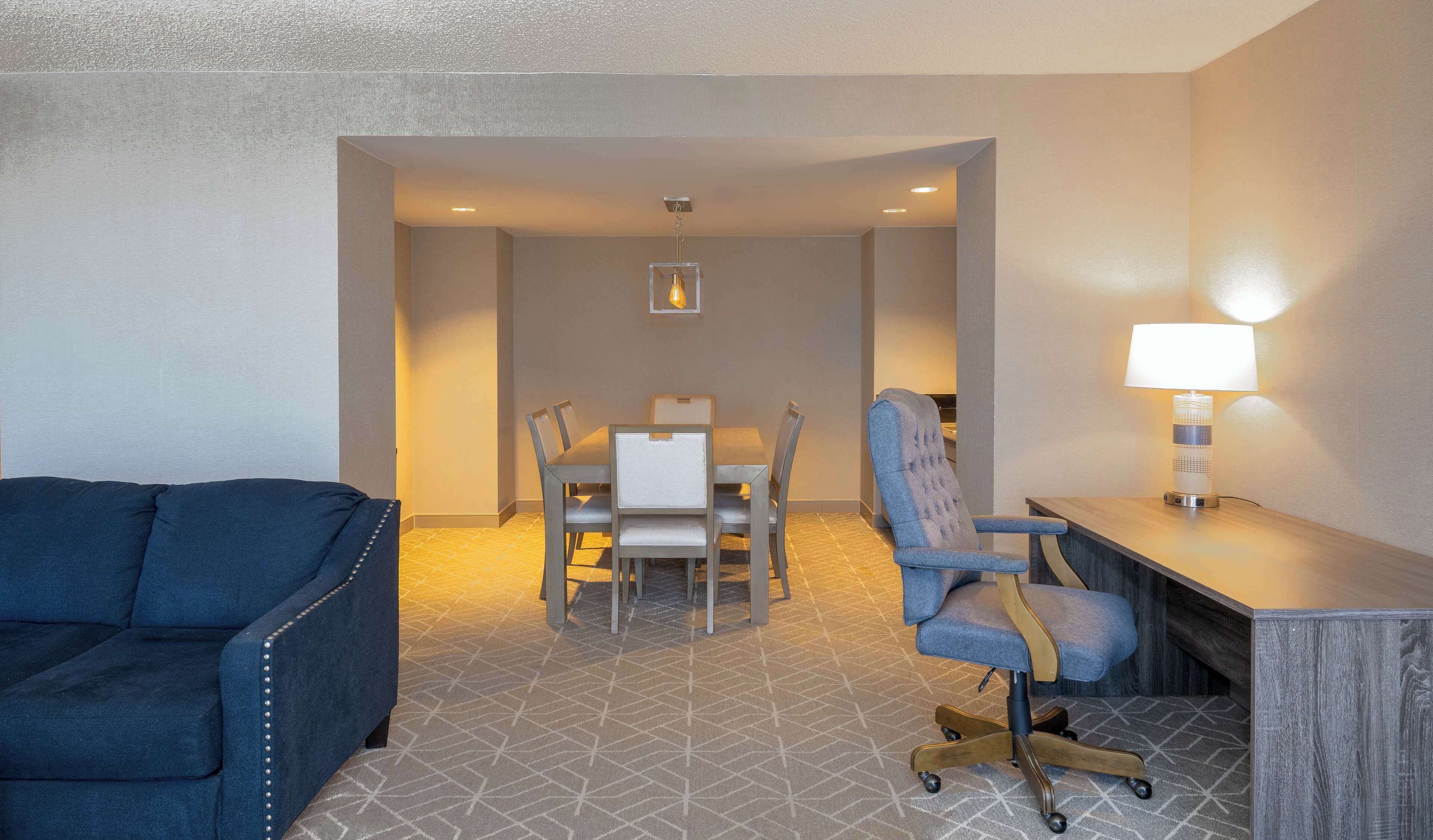A hotel room with a blue sofa, a dining table with chairs, a desk with a lamp, and a patterned carpet.