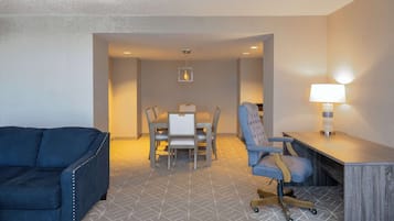 A hotel room with a blue sofa, a dining table with chairs, a desk with a lamp, and a patterned carpet.