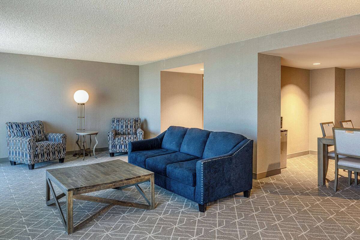 Quarto de hotel com um sofá azul, poltronas estampadas, uma mesa de centro de madeira e uma área de jantar com cadeiras e uma mesa.