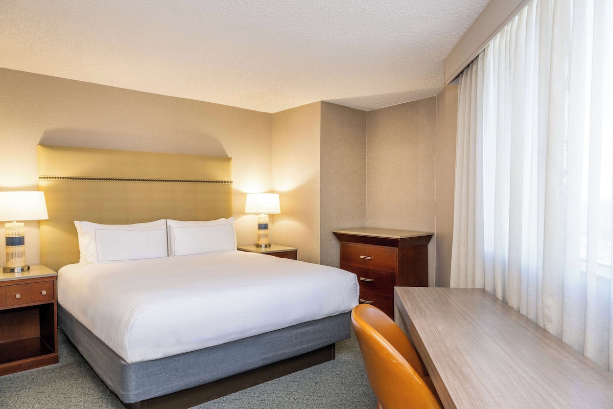Quarto de hotel com uma cama grande, duas mesas de cabeceira com abajures, uma escrivaninha de madeira e uma janela com cortinas.