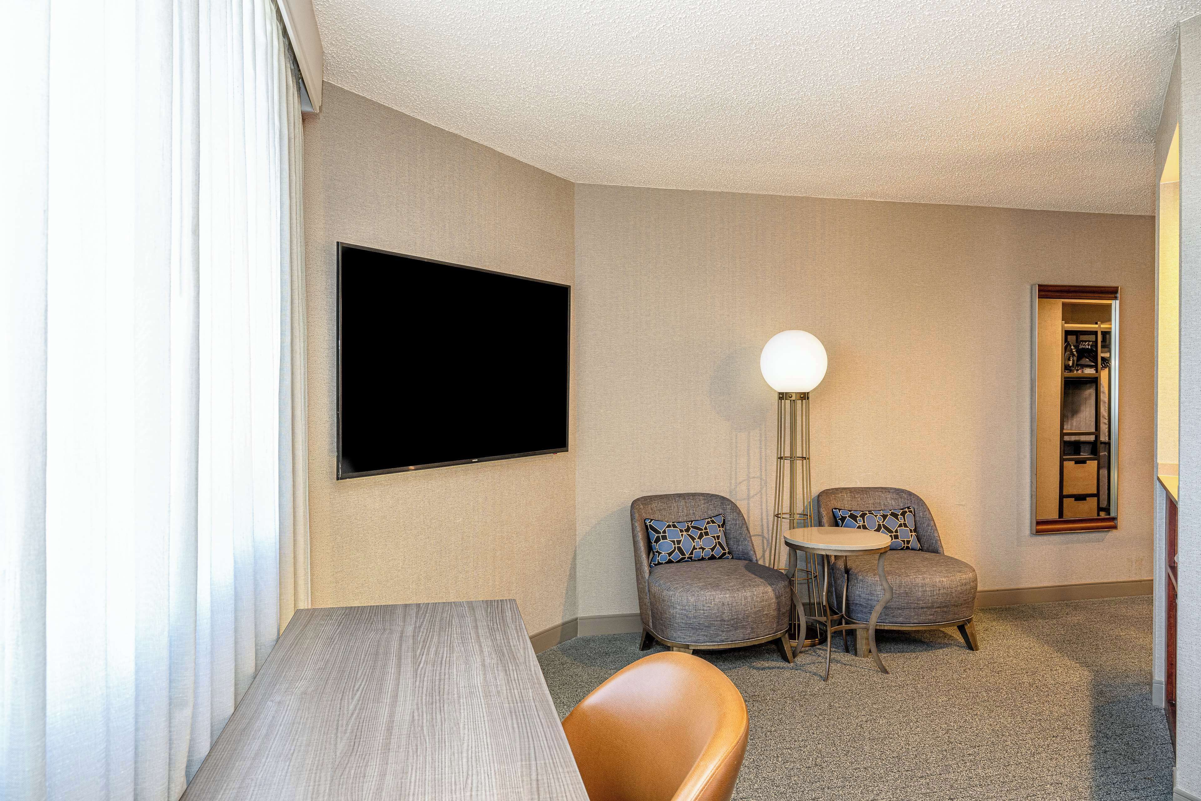 A hotel room with a flat-screen TV mounted on the wall, two armchairs with patterned cushions, a small round table, and a wooden coffee table.