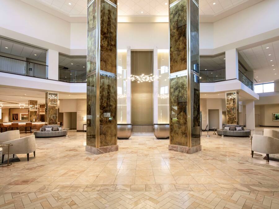 A spacious lobby with a high ceiling, marble flooring, and a central chandelier. There are seating areas with sofas and armchairs.