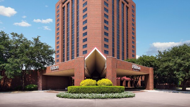 A tall brick building with the Hilton logo, featuring a large entrance canopy and a landscaped area with bushes and flowers. - Hilton Richardson Dallas (Richardson)