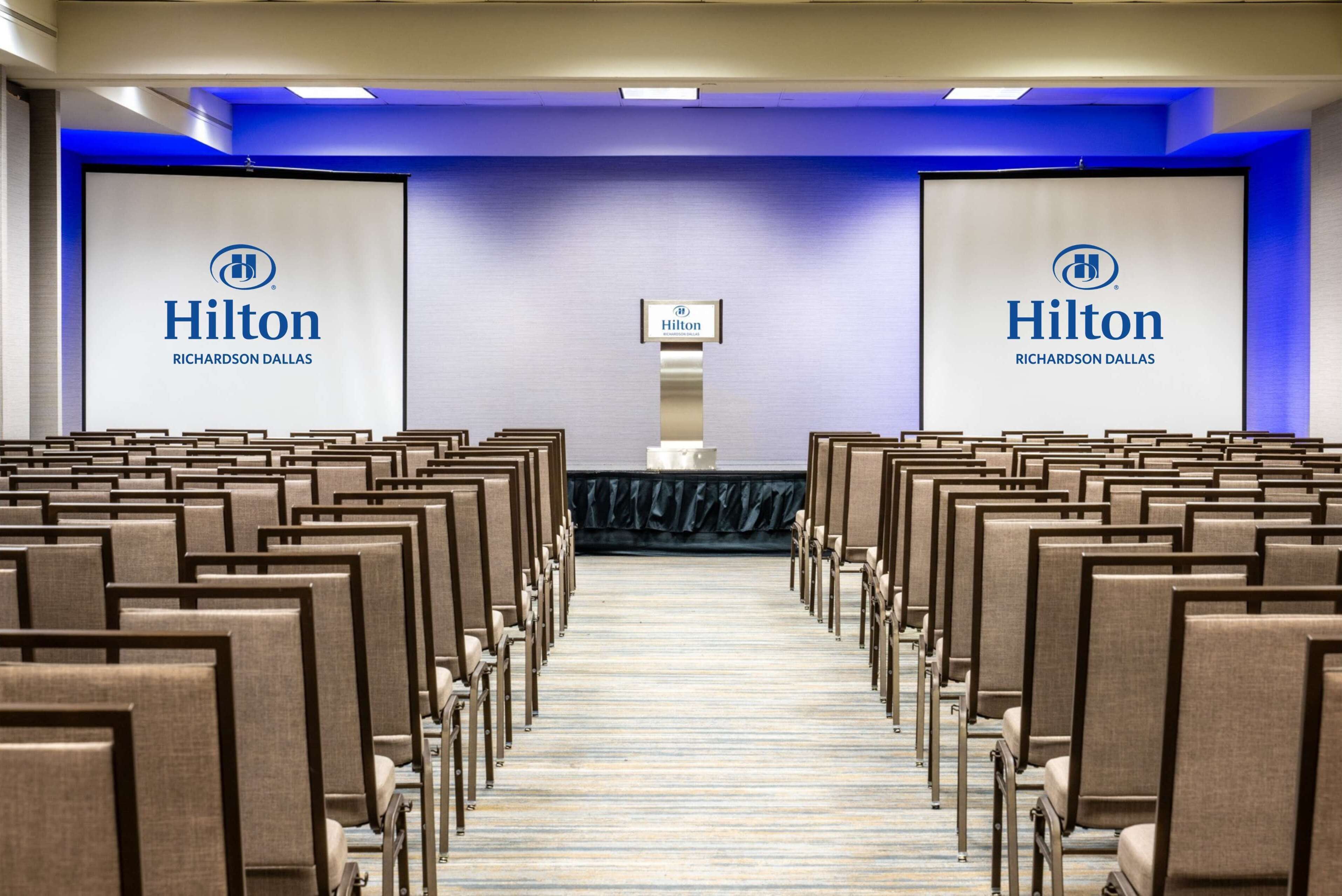 A conference room with rows of chairs and two large screens displaying the Hilton logo.