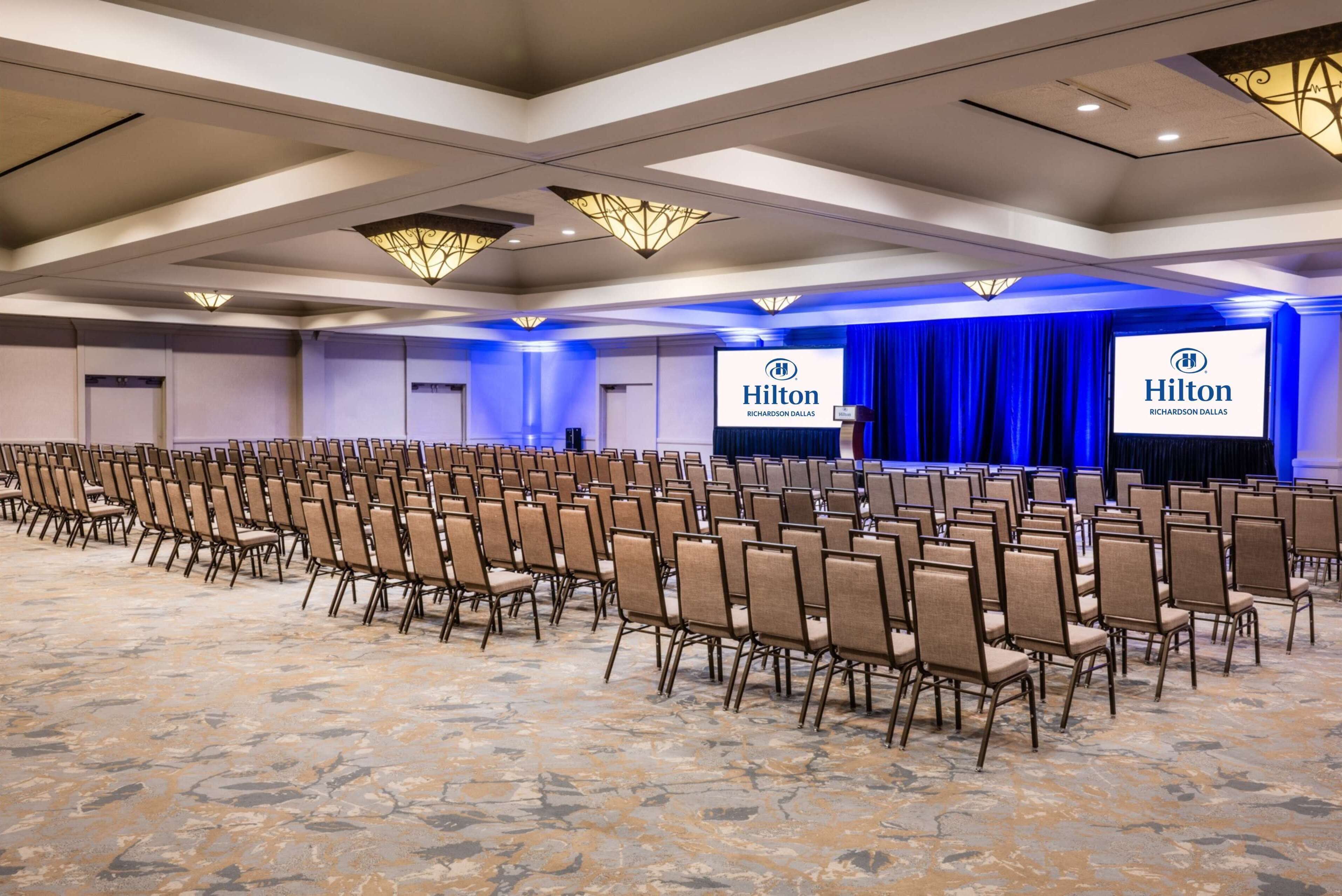 Un salón de conferencias con filas de sillas, un escenario con una pantalla mostrando el logotipo de Hilton, y un telón de fondo azul.