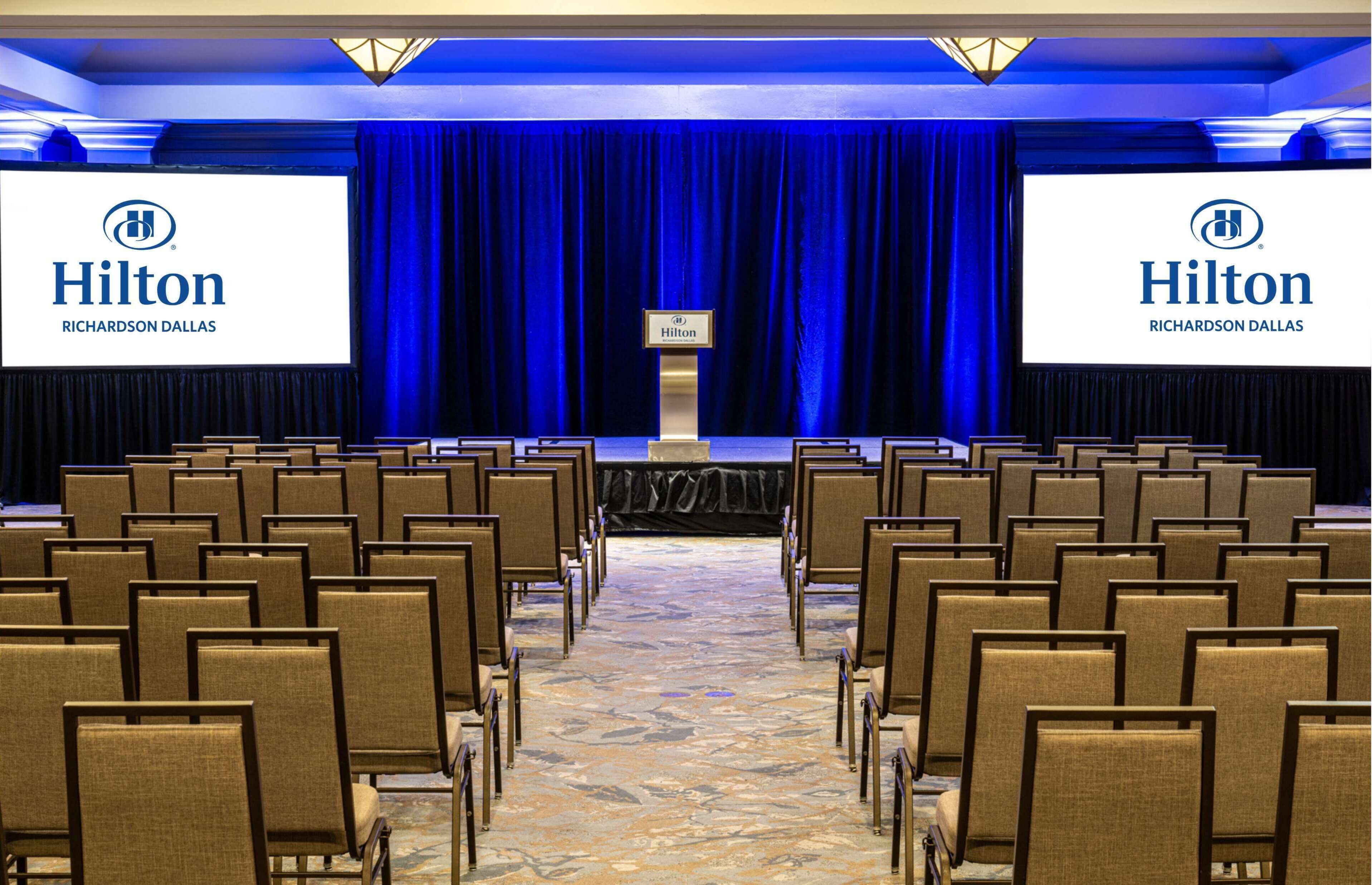 Una sala de conferencias con filas de sillas orientadas hacia un escenario con un telón de fondo azul. Dos pantallas grandes muestran el logo de Hilton Richardson Dallas.