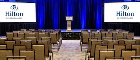 Una sala de conferencias con filas de sillas orientadas hacia un escenario con un telón de fondo azul. Dos pantallas grandes muestran el logo de Hilton Richardson Dallas.