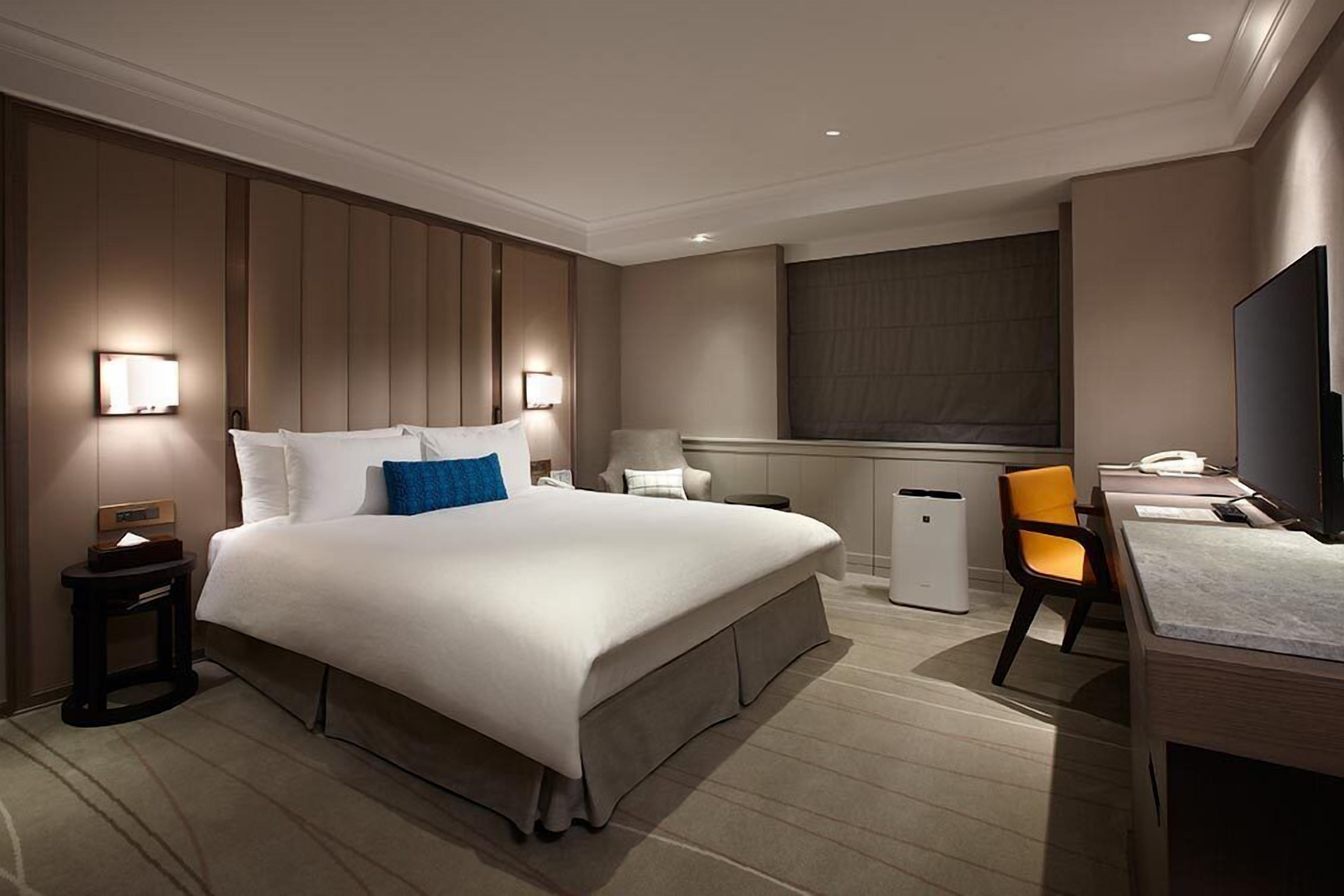 A hotel room with a large bed, bedside tables, a desk, a chair, a television, and a window.