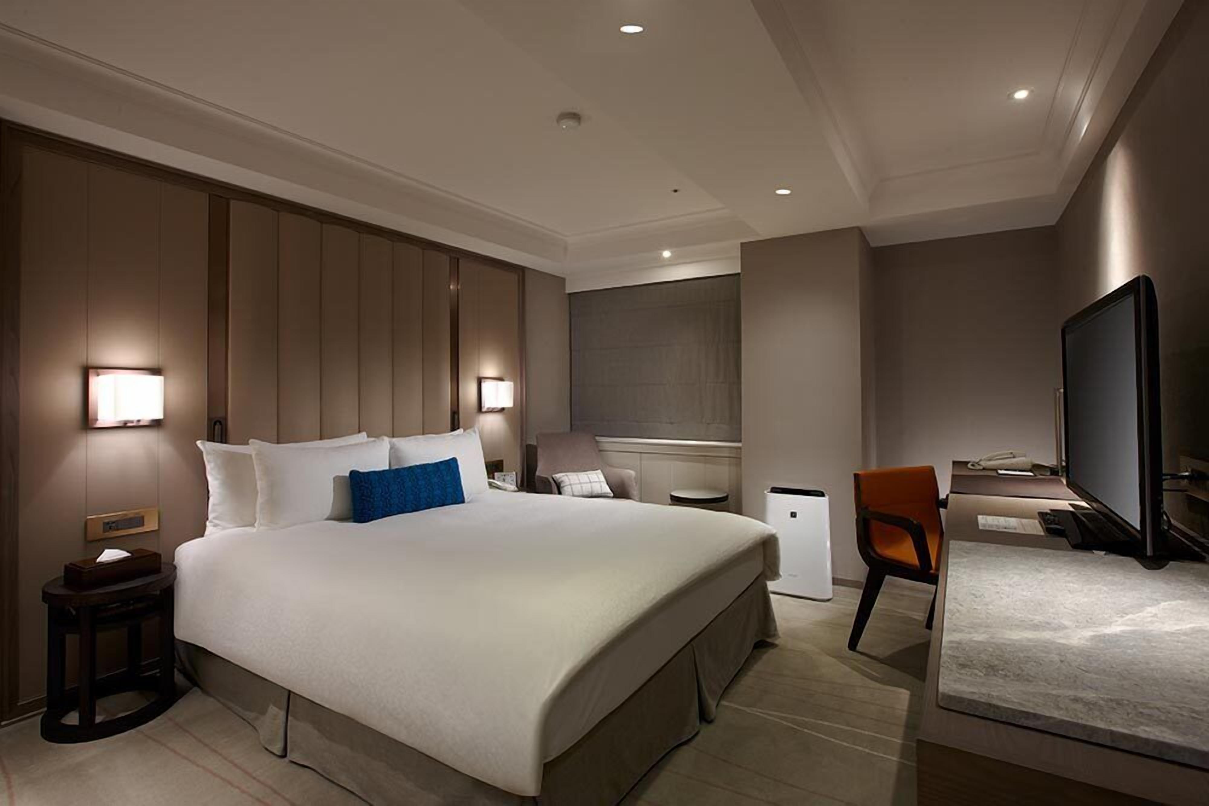 A modern hotel room with a large bed, bedside tables, a desk, a chair, a television, and a small refrigerator.