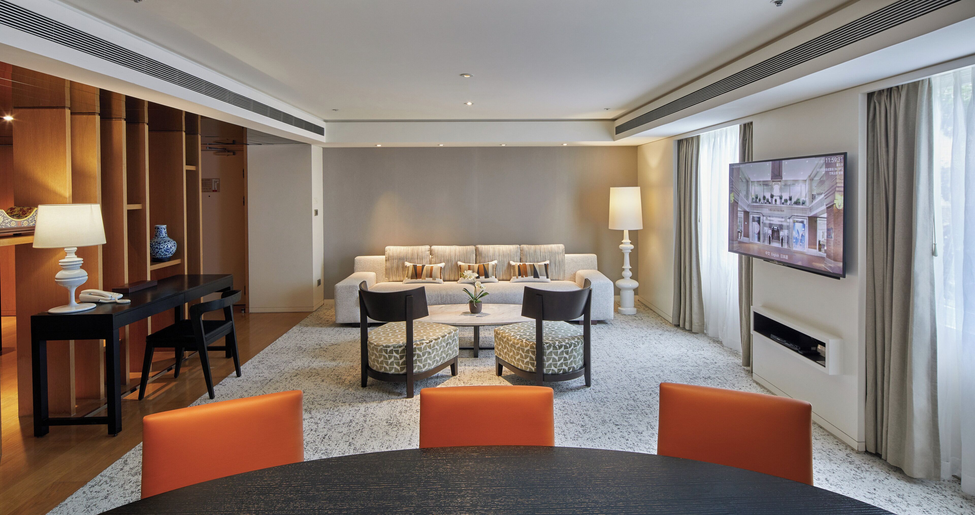 Una habitación de hotel moderna con un conjunto de sofás, una mesa de comedor con sillas, un televisor montado en la pared y un jarrón decorativo.