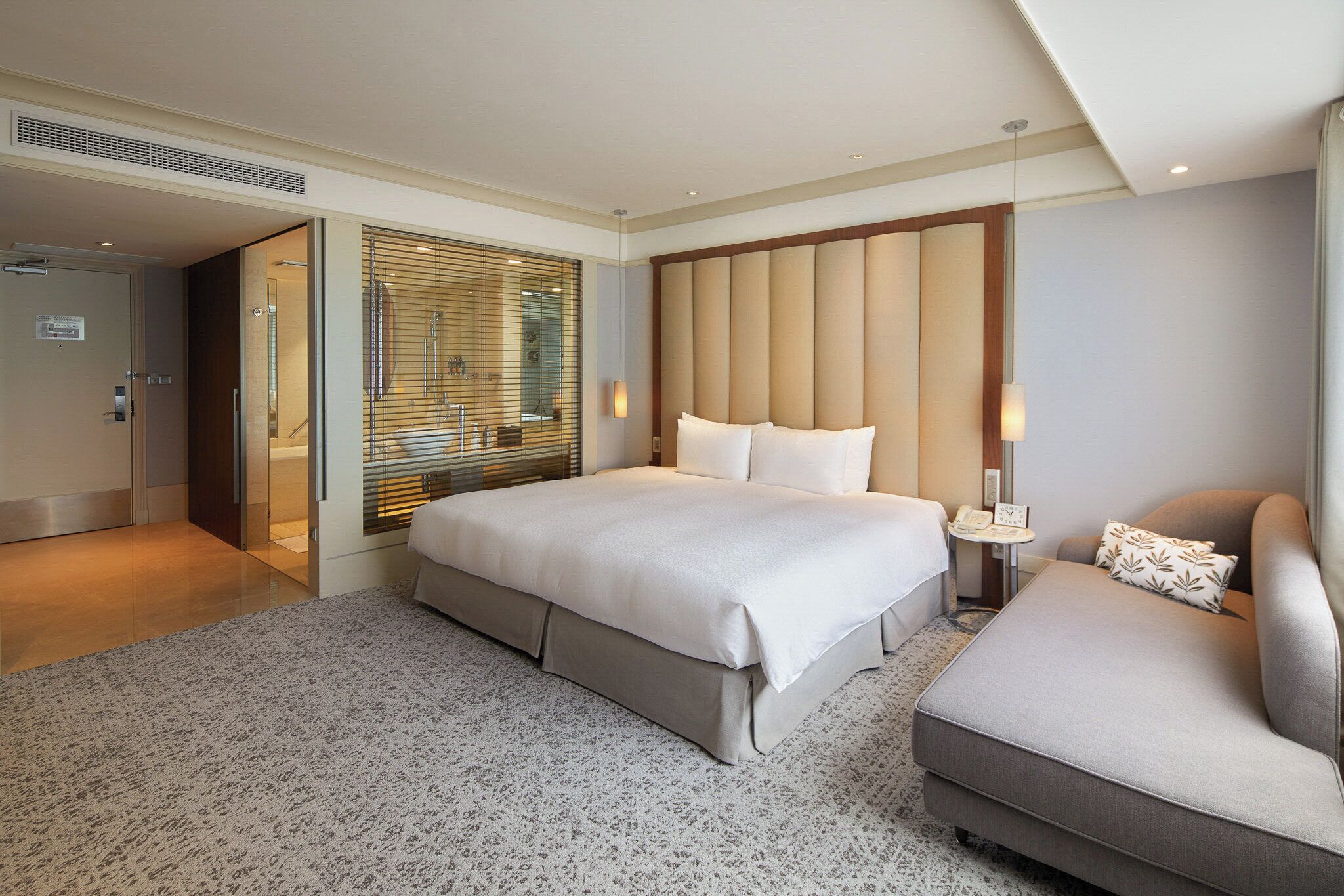 A modern hotel room with a large bed, a sofa, a small table with a lamp, and a sliding door leading to a bathroom.
