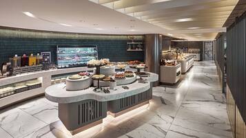 A modern hotel lobby with a marble floor, a central buffet area with various food items, and a large screen displaying a cityscape.