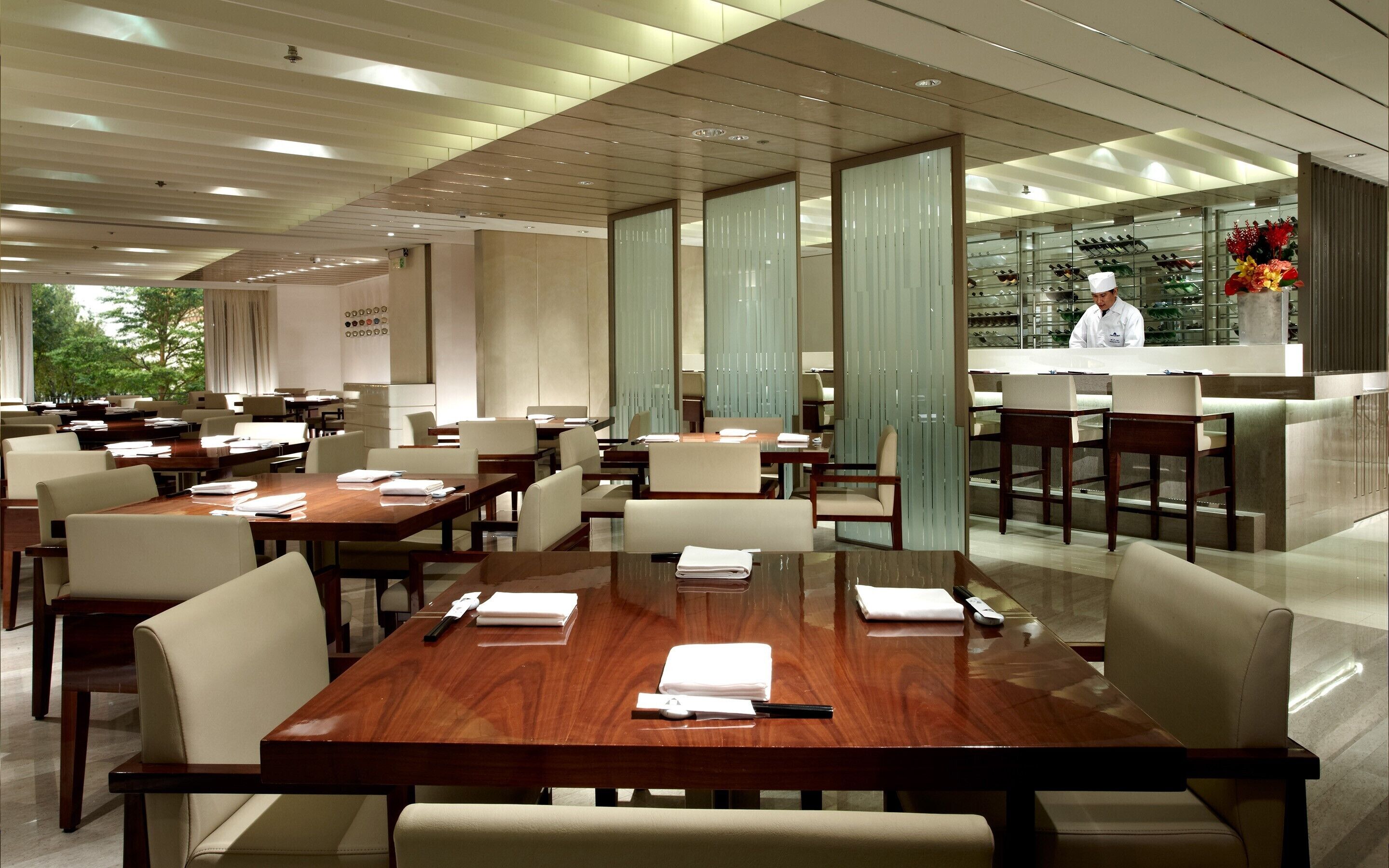A modern conference room with wooden tables, white chairs, and a glass partition separating the dining area from the kitchen.