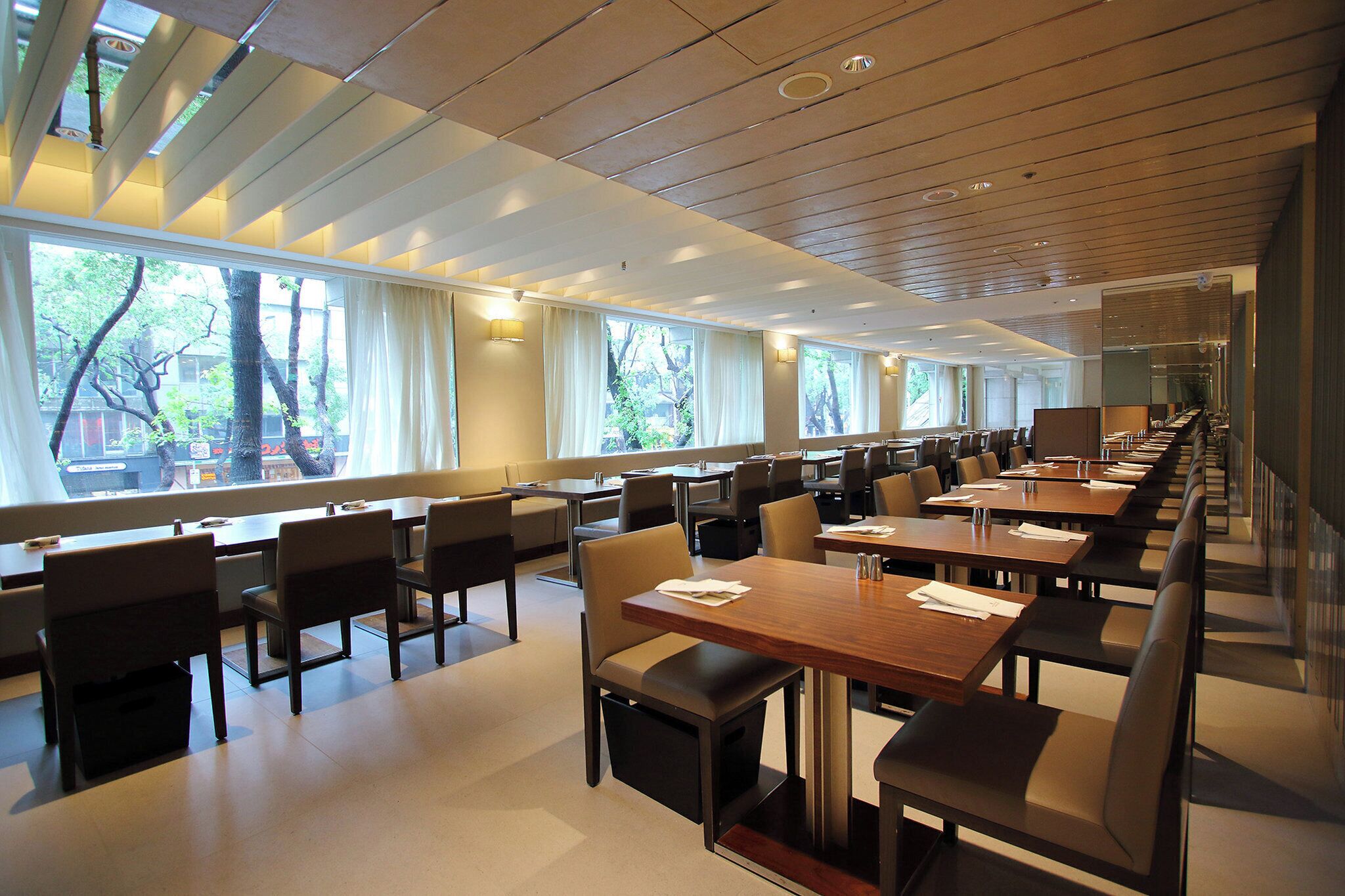 A modern dining area with long tables and chairs, large windows, and a wooden ceiling.