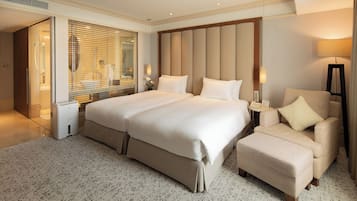 A spacious hotel room with a large bed, a seating area with a chair and ottoman, a bedside table with a lamp, and a view through the window blinds.