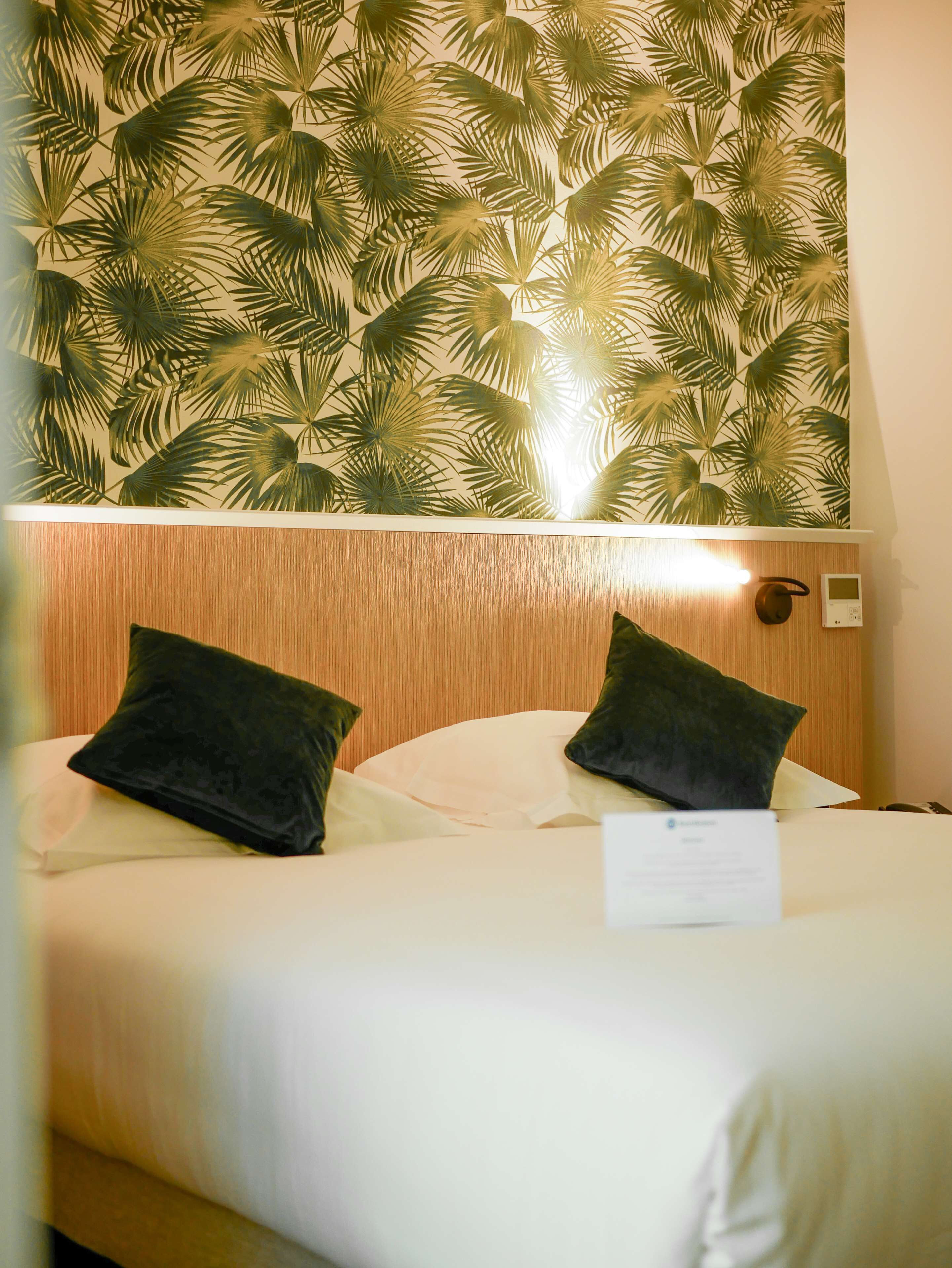 A hotel room with a bed, two dark pillows, a wooden headboard, a wall with a palm leaf pattern, and a thermostat on the wall.