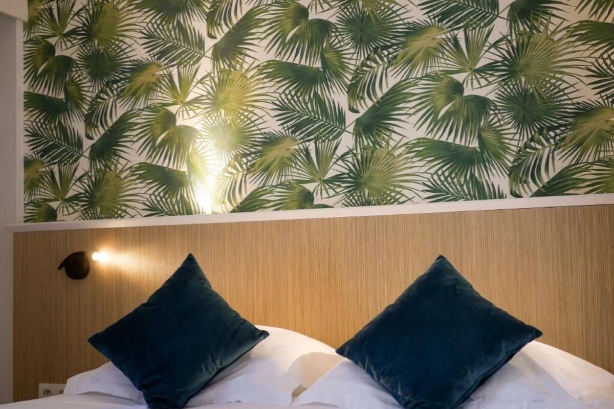 Une chambre d’hôtel avec un lit, deux oreillers foncés, un mur orné d’un motif de feuilles de palmier et un luminaire placé au-dessus du lit.