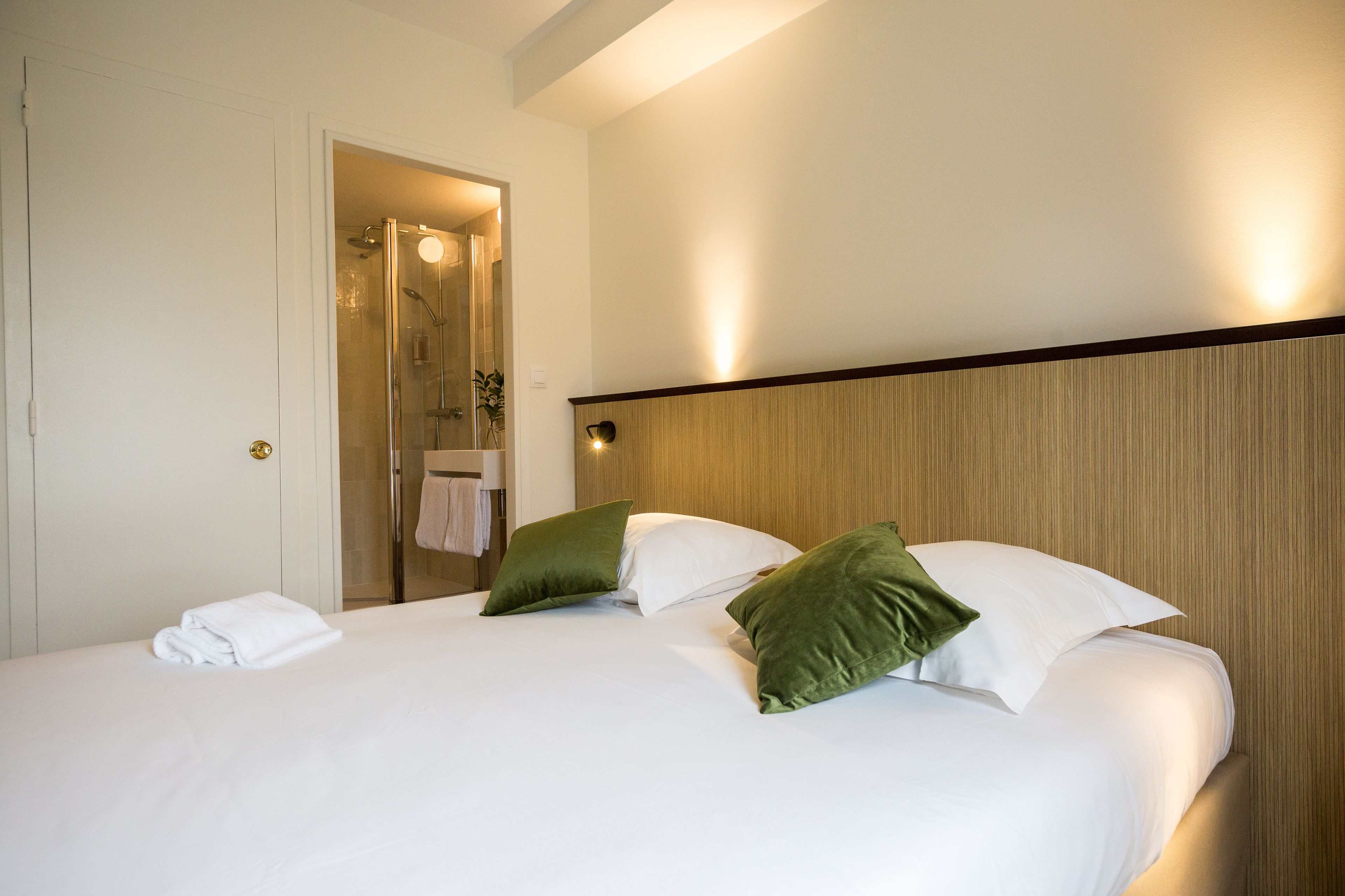 A hotel room with a bed, green pillows, a white blanket, and a shower area visible through an open door.