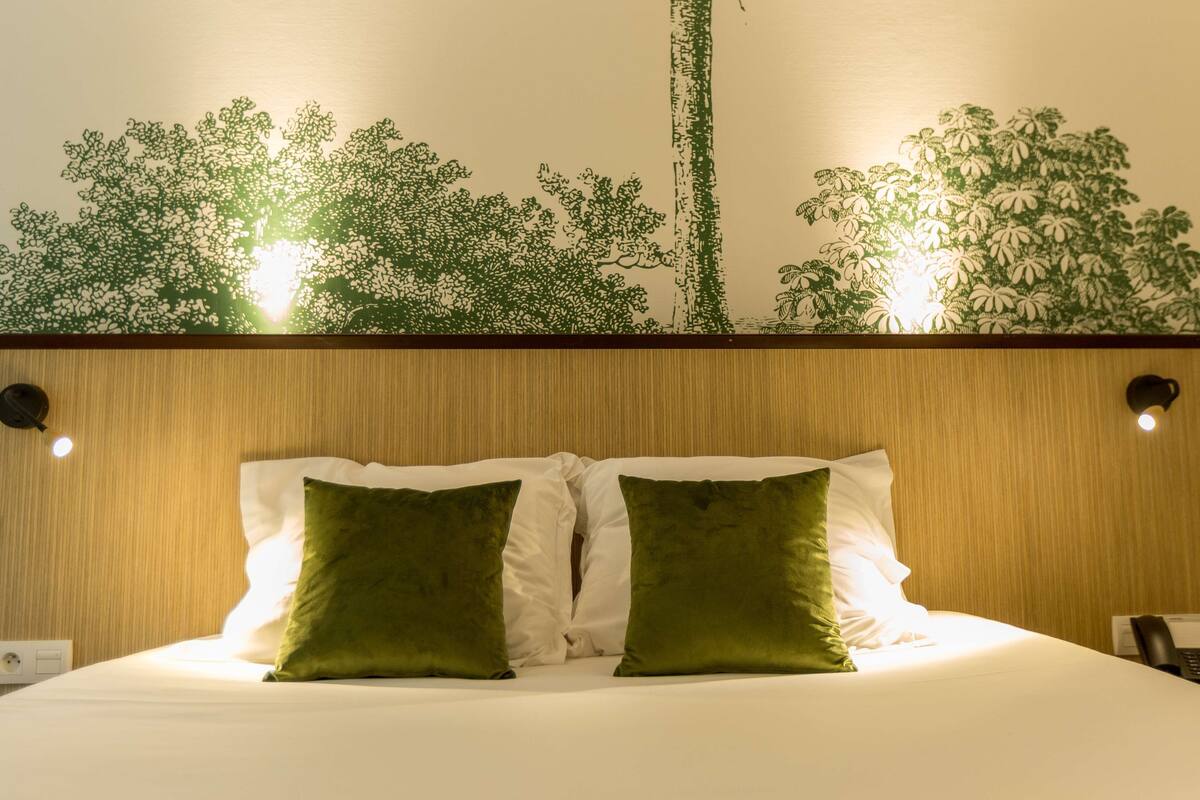 Un lit bien fait, avec deux oreillers verts, une tête de lit en bois et un mur orné d’un motif végétal vert.