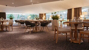 Un vaste espace dédié à la salle à manger, avec de nombreuses tables et chaises, de grandes fenêtres dotées de rideaux et des plantes en pot.