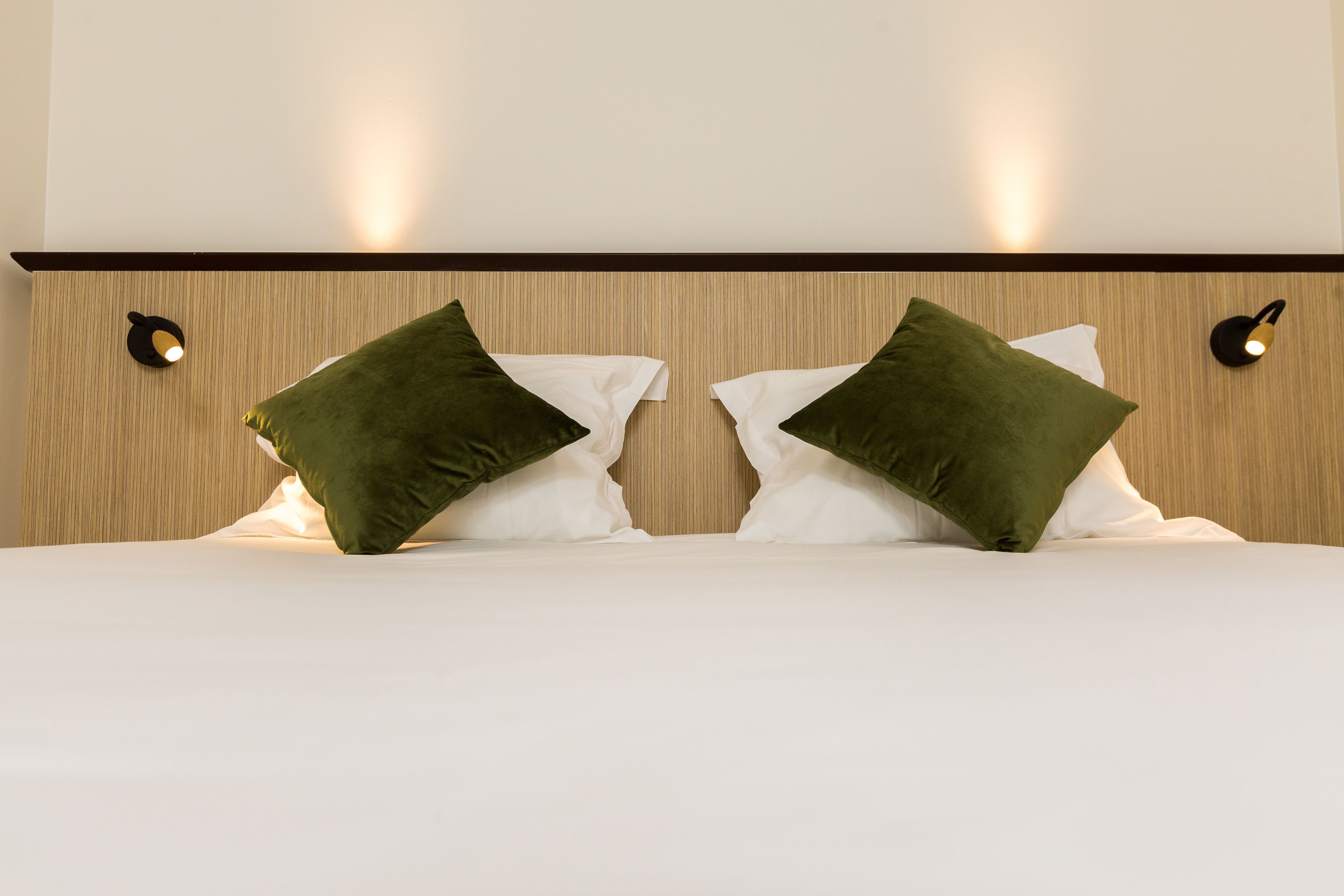 A neatly made bed with white linens and two green pillows, a wooden headboard with vertical lines, and two wall-mounted light fixtures on either side.