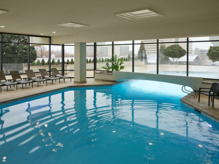 An indoor swimming pool with clear views of the outside through large windows, lounge chairs, and a potted plant.