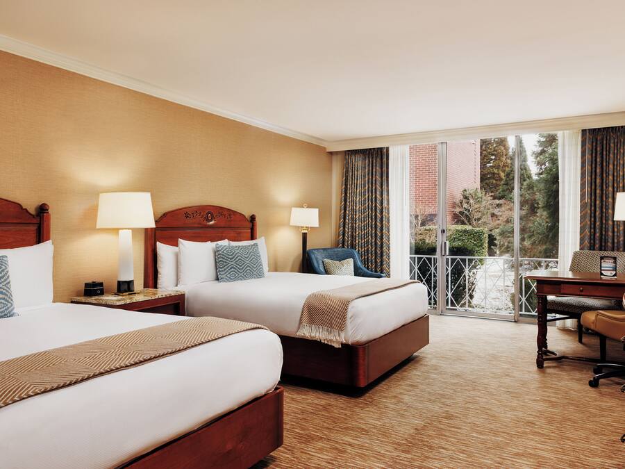 A hotel room with two beds, a desk, a chair, and a view of the outdoors.