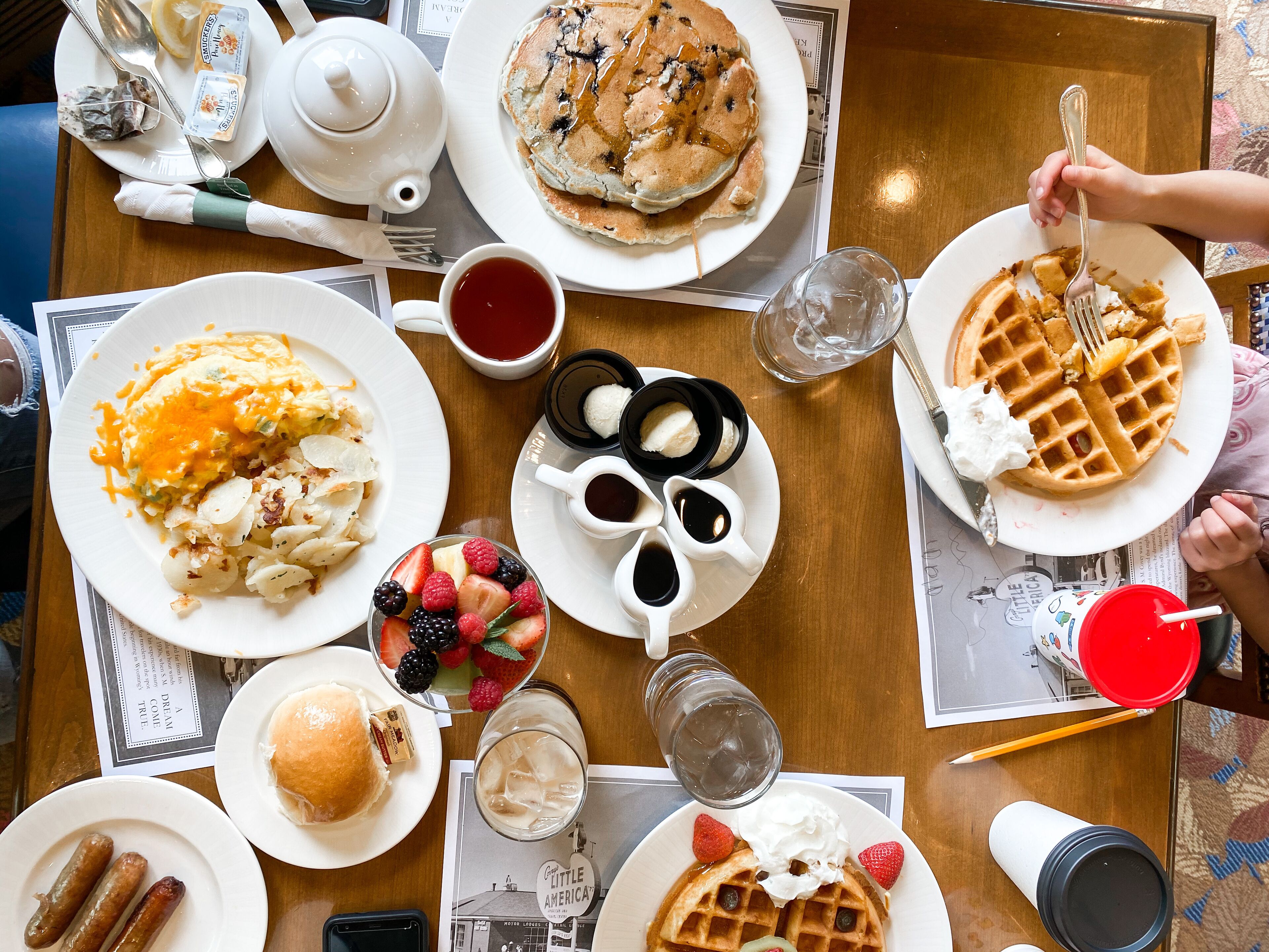 Une table de petit-déjeuner avec divers plats, notamment des crêpes, des gaufres, des saucisses et un saladier de fruits.