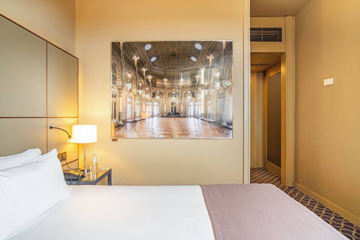 A hotel room with a large bed, a bedside table with a lamp, and a framed picture of a grand interior.