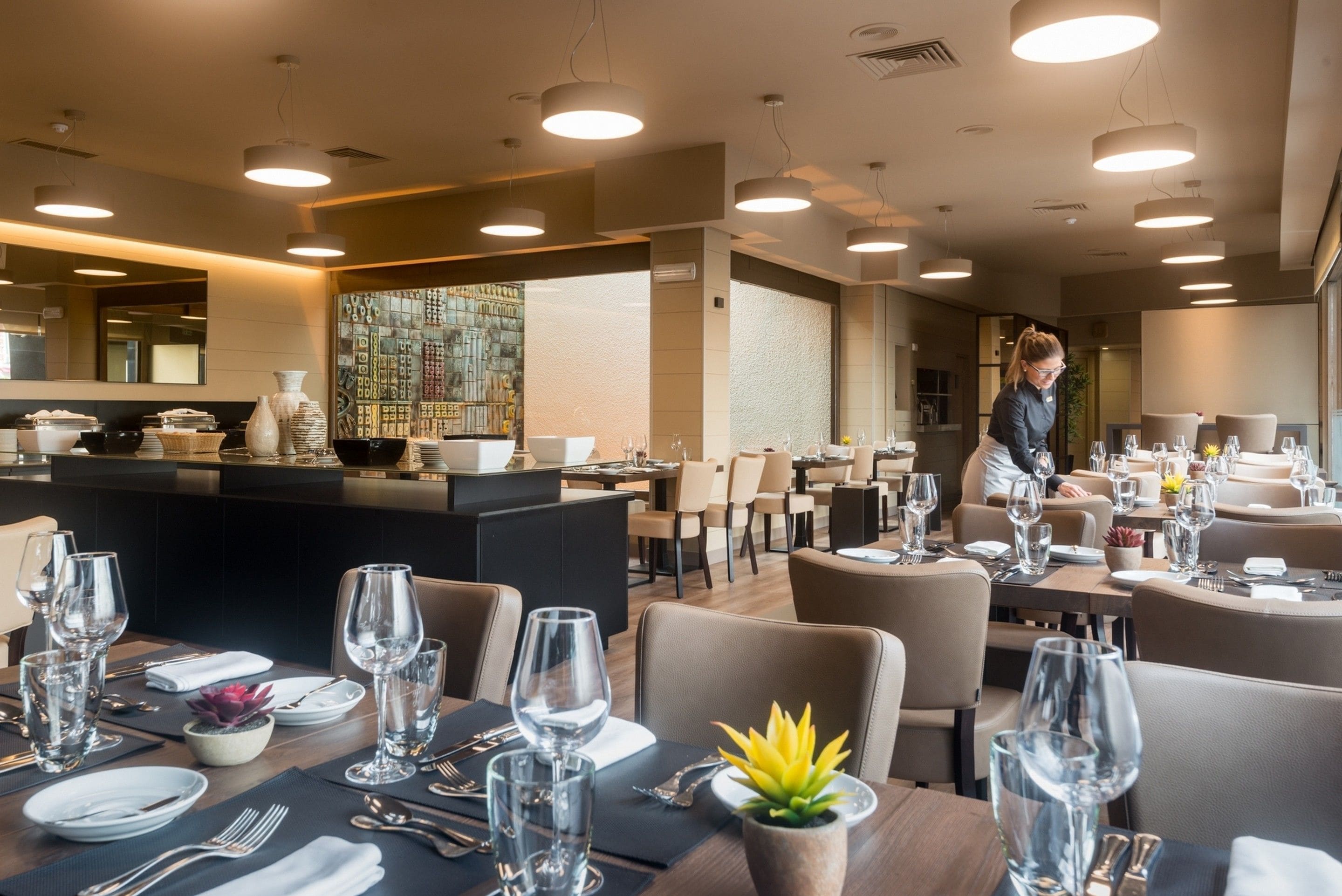 Un restaurant moderne doté de nombreuses tables, chacune mise avec des verres à vin, des assiettes et des couverts. On aperçoit un bar à l’arrière-plan, avec un barman.